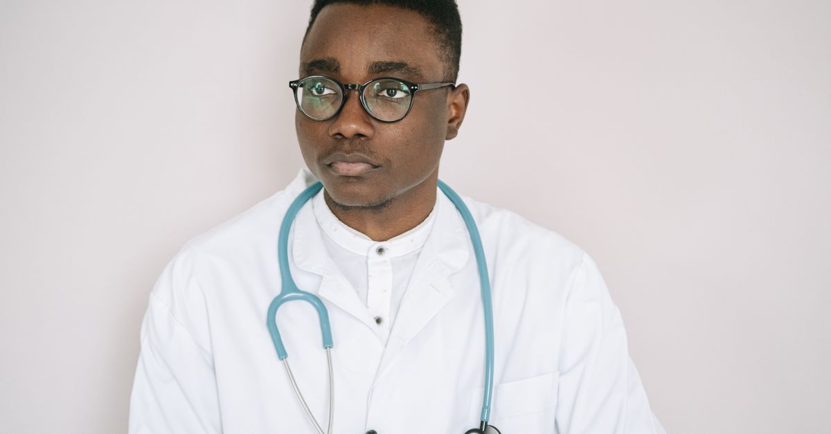 Médico de jaleco branco em postura profissional, consultório ao fundo