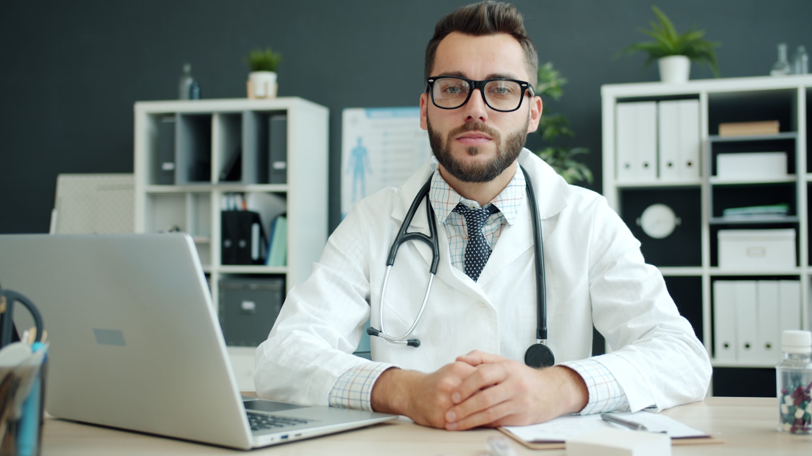 Médico jovem em consultório próprio moderno, sentado à mesa com laptop, esperando o primeiro paciente chegar